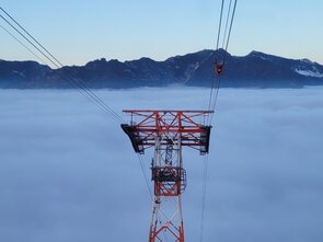 Seilbahnstütze ragt über Wolken, mit Bergen im Hintergrund.