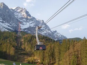 Seilbahn fährt über einen Wald mit schneebedeckten Bergen im Hintergrund.