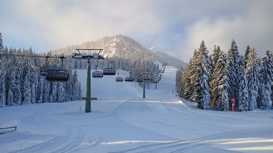 Snow-covered slope with a chairlift and pine trees in the background.