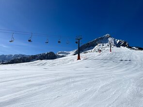 Wide snowy slope with a chairlift and mountains in the background.