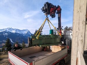 A crane is lifting a large green machine over a truck in the Alps.