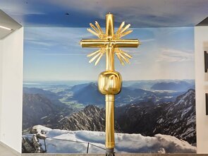 A golden cross in front of an impressive mountain landscape.