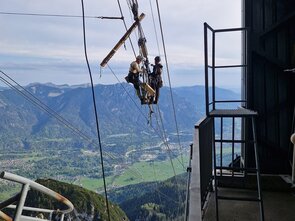 Zwei Personen mit Helm hängen mit einem Klettergurt an den Seilen einer Seilbahn und verrichten dort Arbeiten. 