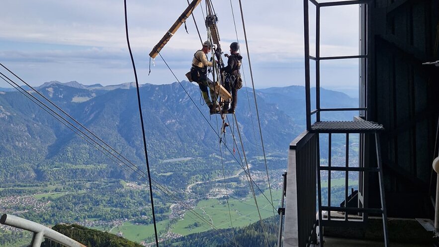 Zwei Personen mit Helm hängen mit einem Klettergurt an den Seilen einer Seilbahn und verrichten dort Arbeiten. 