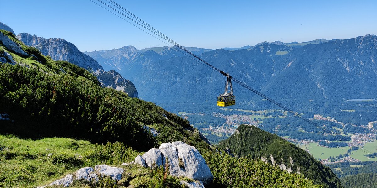 Eine Seilbahn schwebt über grüne Wiesen und Berglandschaft.