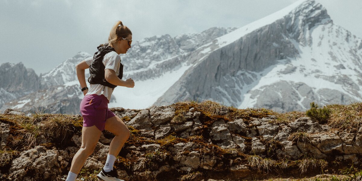 A trail runner runs uphill on a path in front of the snow-covered Alpspitze in summer. 