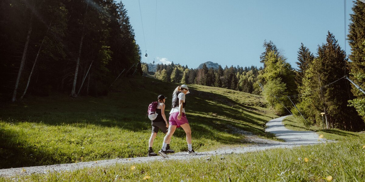 Zwei Frauen gehen auf einem breiten Wanderweg durch grüne Wiesen unterhalb der Seilbahn.