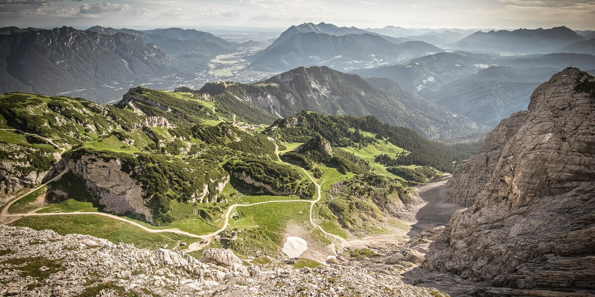 Aussicht auf eine Berglandschaft mit grünen Wiesen, schroffen Felsen und verschiedenen Wanderwegen. 
