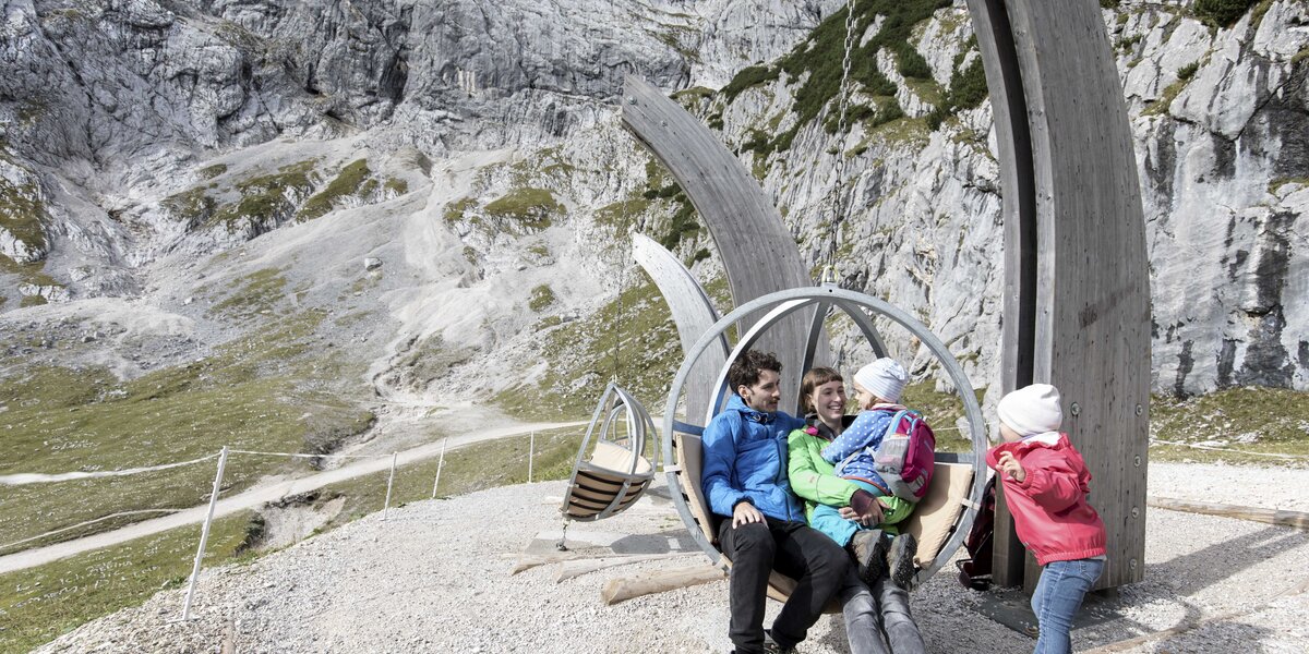 A family sits in spherical swings in front of a rugged rock face.