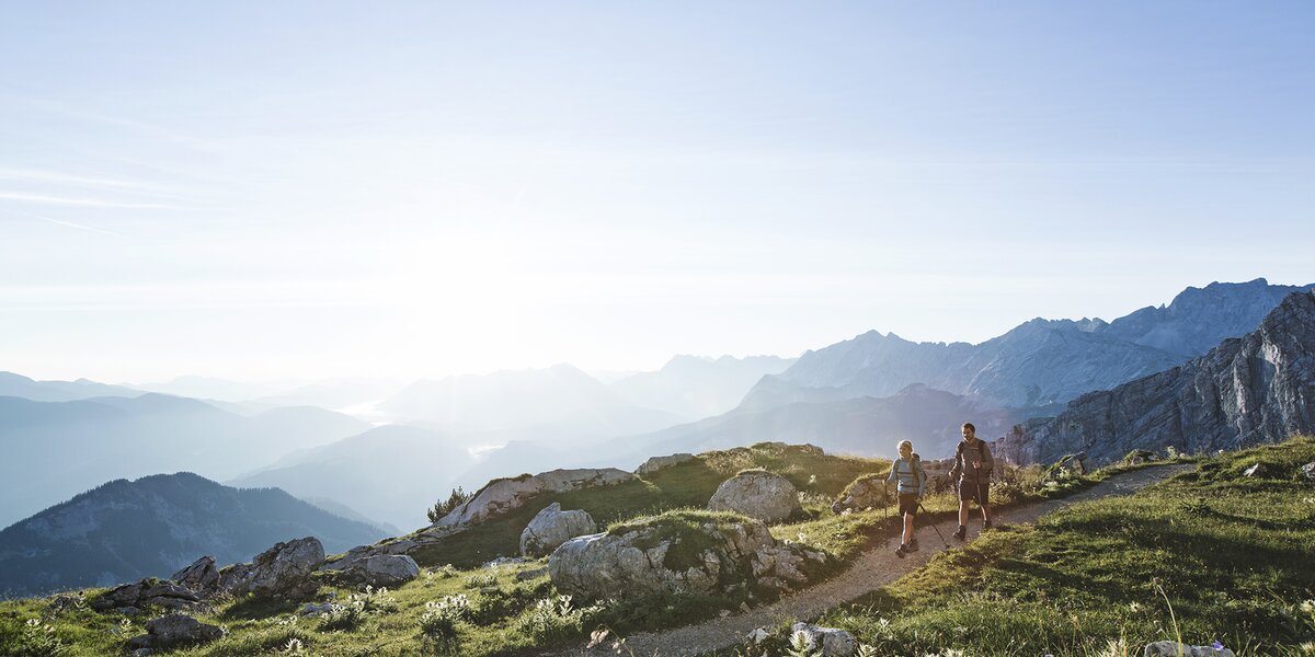 Ein Mann und eine Frau gehen in der Morgensonne auf einem Wanderweg über eine alpine Wiese.