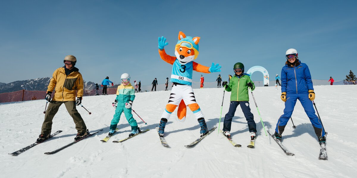 Two adults and two children are skiing in the children's area with the mascot Ixi, a fox dressed in ski racing gear. Ixi waves cheerfully at the camera.