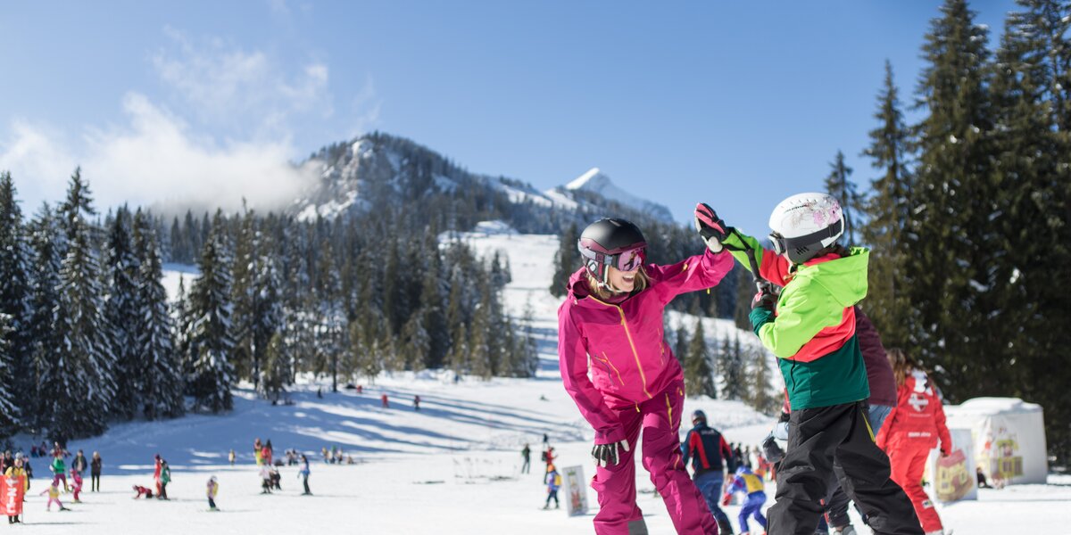 A woman and a child on skis high-five each other. In the background, you can see numerous visitors to the ski resort and snow-covered mountains. 