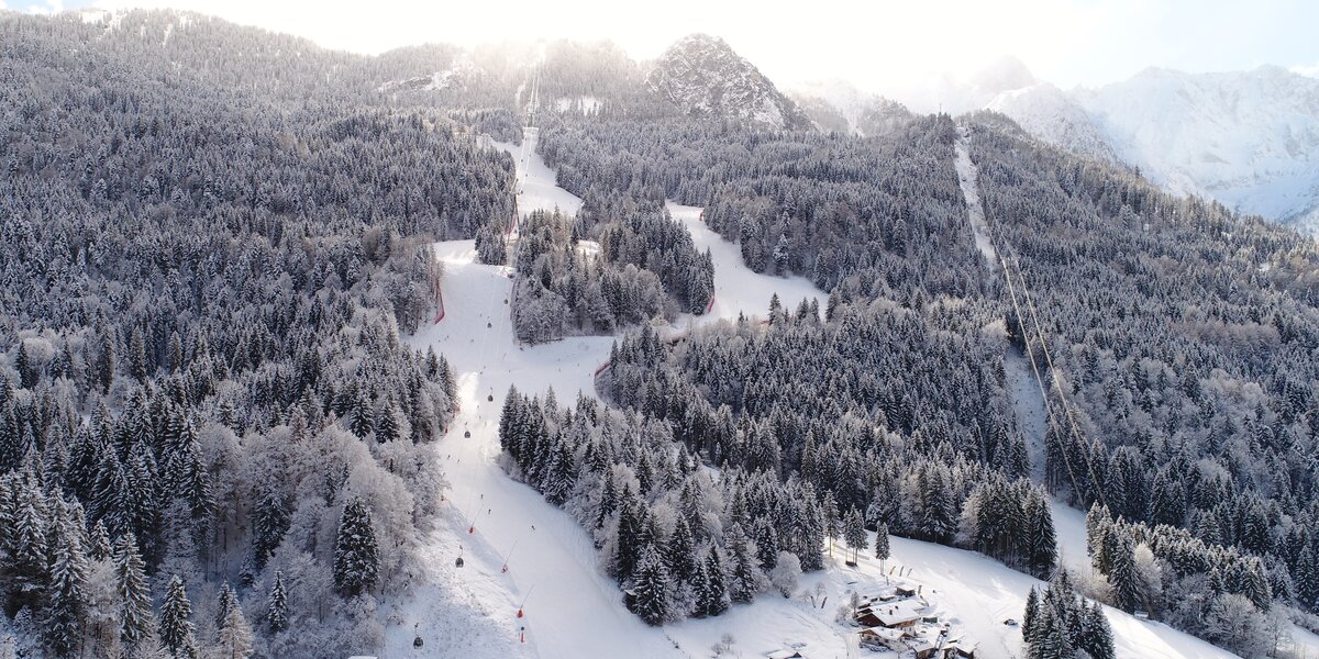 Drohnenaufnahme der Kandahar-Abfahrt mit Kreuzeck- und Alpspitzbahn vor schneebedeckten Wäldern.