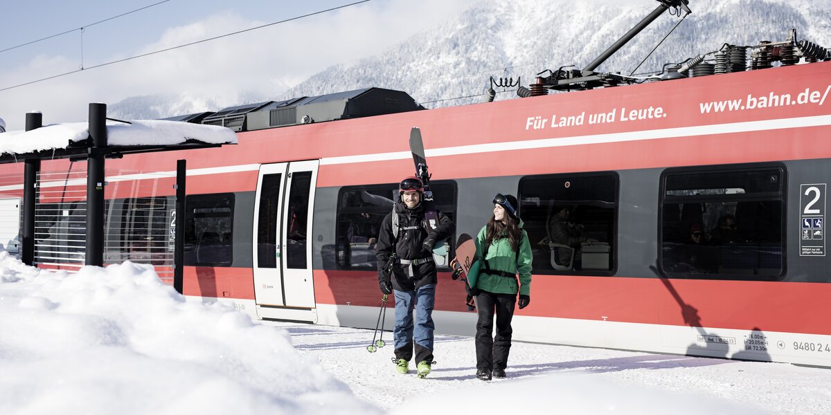 A skier and a snowboarder are walking on a snow-covered platform where a red DB train is standing. 