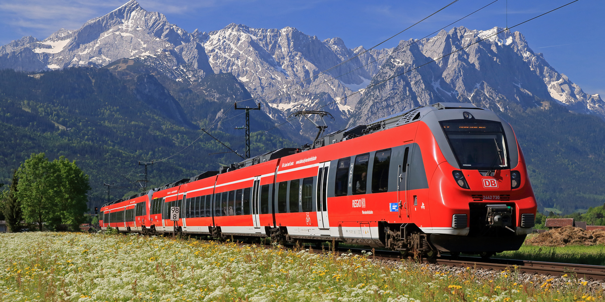 Ein roter Zug der DB fährt durch eine blühende Landschaft mit Alpen im Hintergrund.
