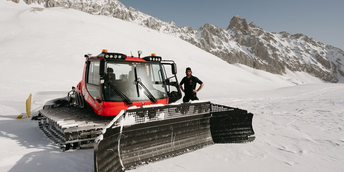 Ein Mitarbeiter steht neben einer Pistenraupe im Schnee vor einer Berglandschaft. 