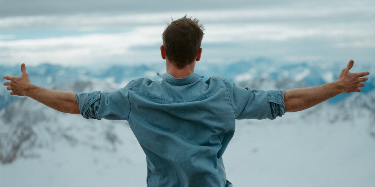 A man with rolled-up sleeves and arms spread wide stands with his back to the camera in front of a snowy mountain panorama. 