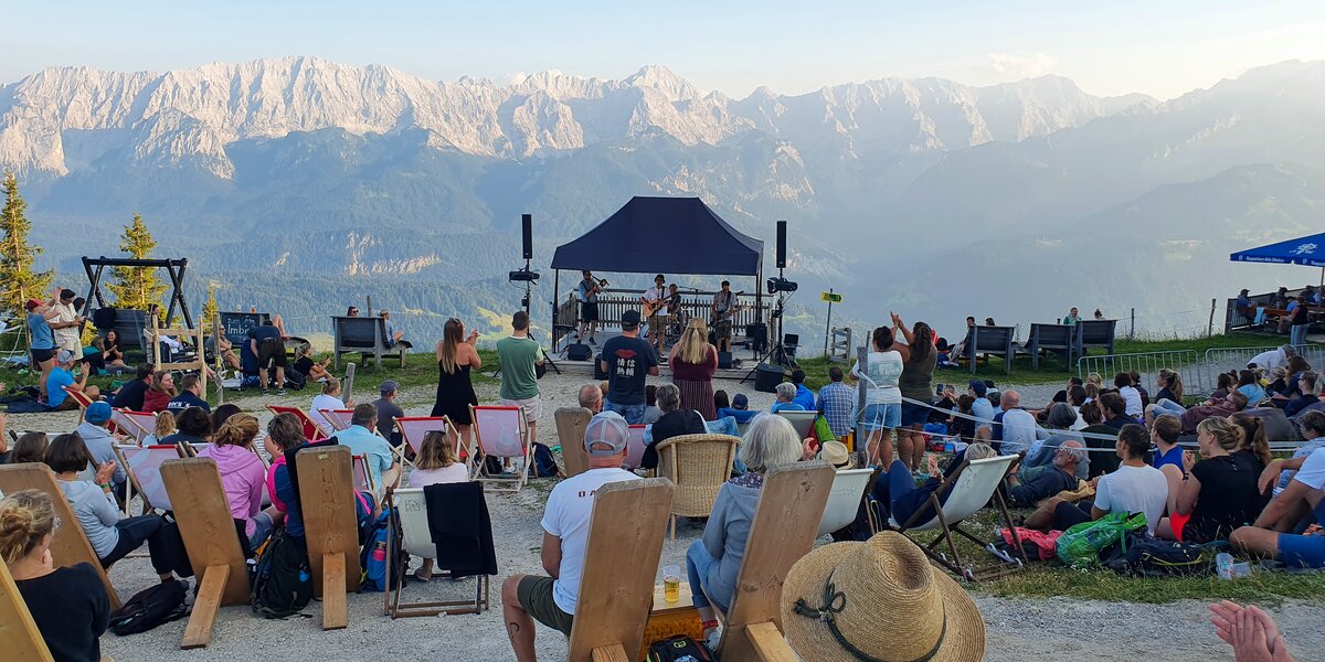 Menschen hören bei einem Konzert auf dem Berg unter freiem Himmel zu und blicken auf das beeindruckende Alpenpanorama hinter der Bühne.