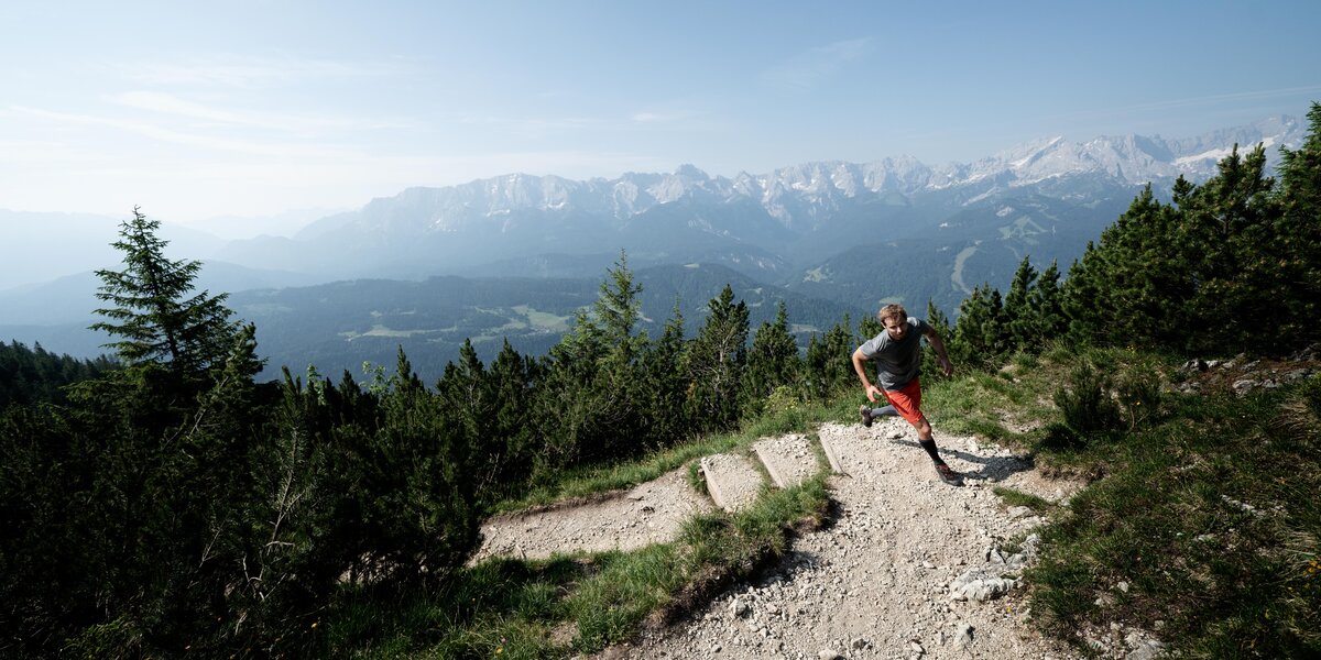 A man jogs up a narrow path in front of an impressive mountain panorama.