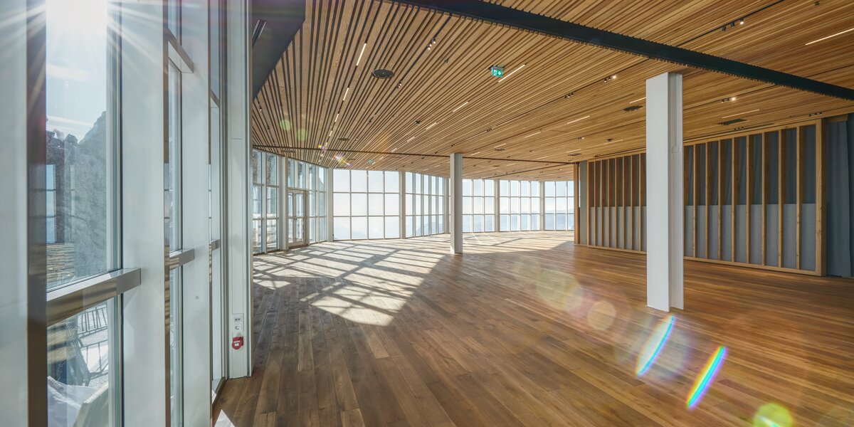 Luftiger Raum mit Holzfußboden und großen Fensterfronten, viel Tageslicht.