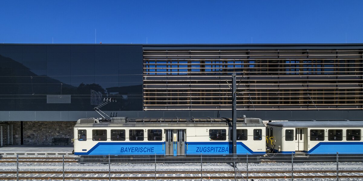 Ein Zug der Bayerischen Zugspitzbahn vor einem modernen Bahnhofsgebäude.