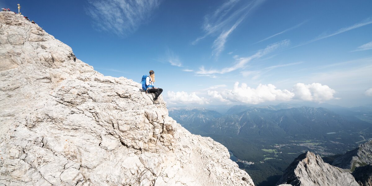Ein Bergsteigern sitzt allein einige Meter unterhalb des Zugspitz-Gipfelkreuzes und genießt die Aussicht. 
