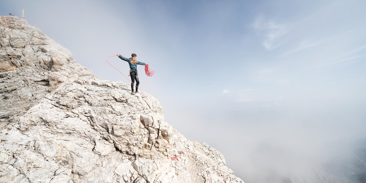 Eine Frau in Kletterkleidung steht auf einem felsigen Gipfel und wickelt ein Kletterseil auf.