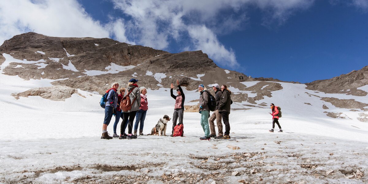Mehrere Personen mit Rucksäcken und sportlicher Kleidung stehen im Sommer auf dem Gletscher auf der Zugspitze und hören der Bergführerin zu.